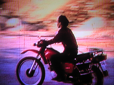 A woman rides a motorcycle against a sunset lit sky