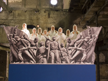 A chorus in white boiler suits stand behind a concrete frieze of Russian workers