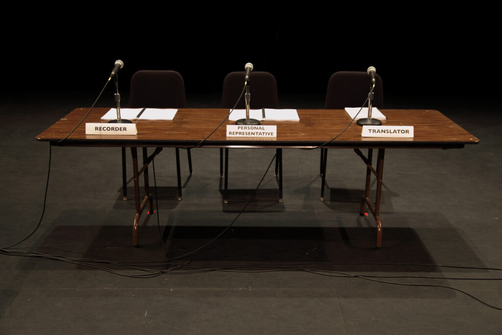 A table with three microphones, chairs and named placards. The seats are empty