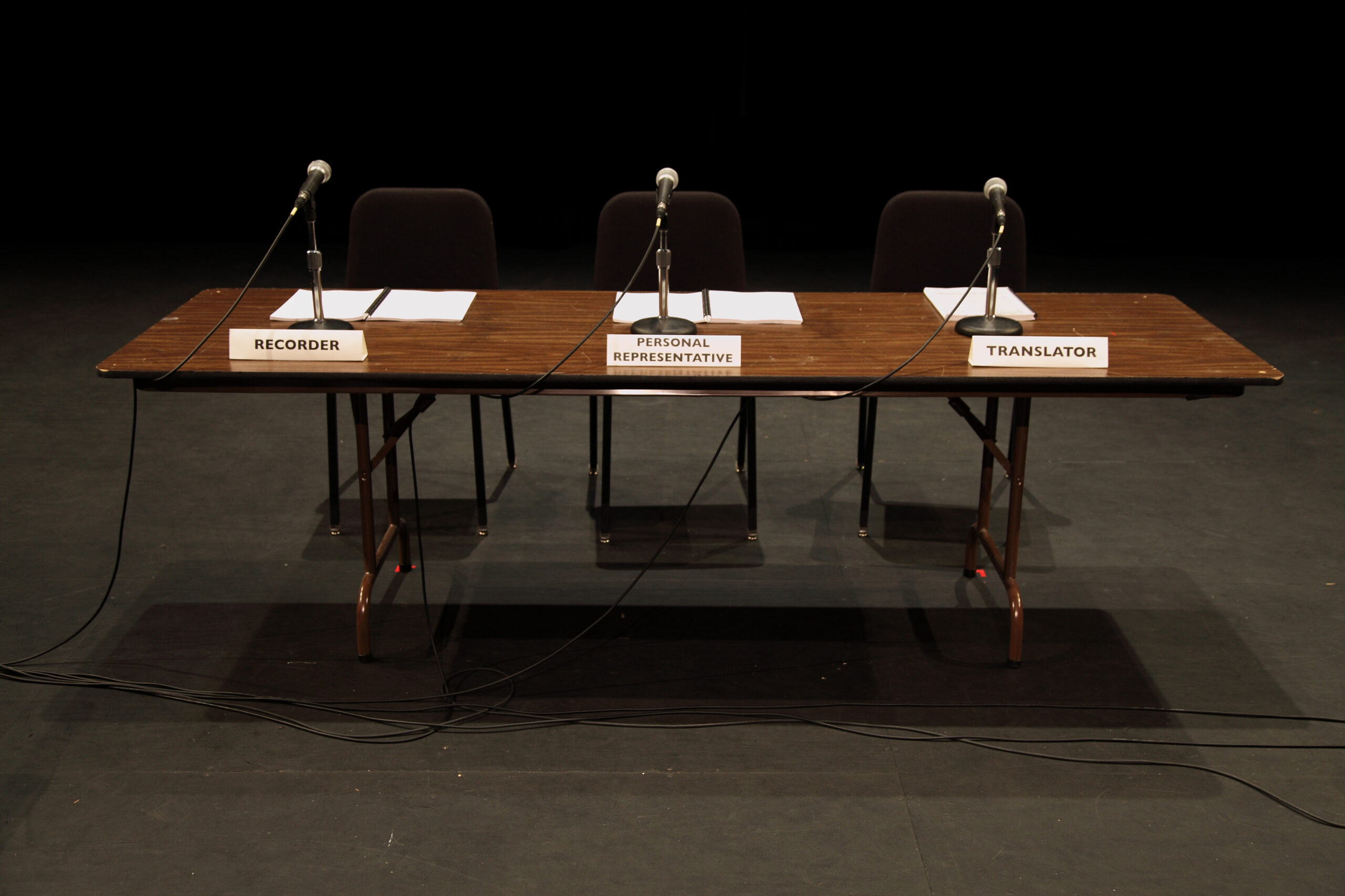 A table with three microphones, chairs and named placards. The seats are empty