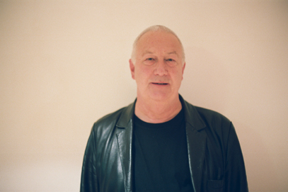 John Tilbury, in a leather jacket, stands against a neutral wall