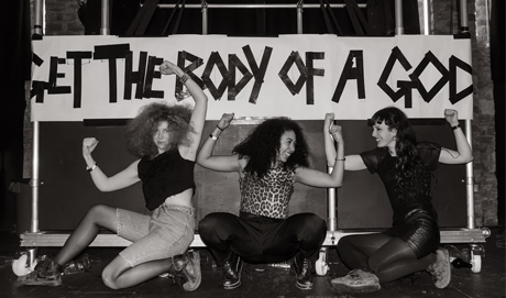 Three women pose in front of a home made banner "Get the Body of a God"