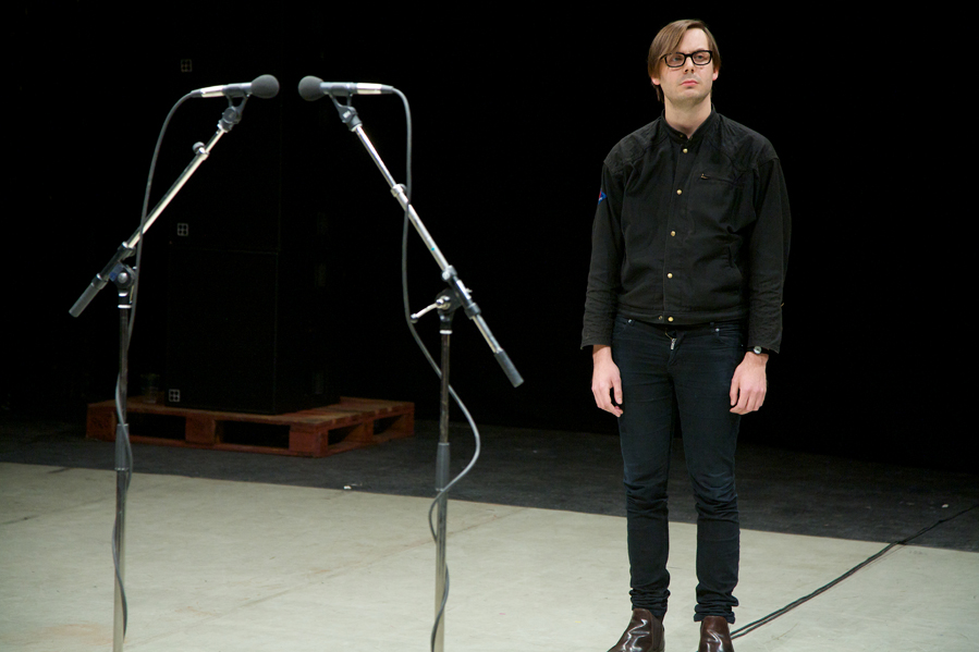 Man in Black clothes standing next to two microphones, staring out