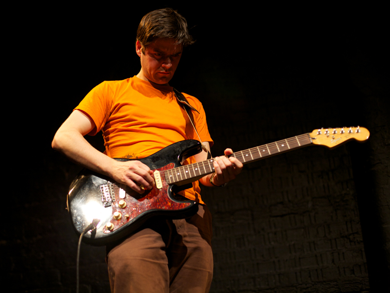 A man in an orange t-shirt plays a red guitar