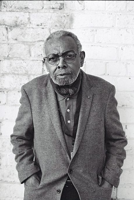 A portrait of Amiri Baraka in front of a brick wall he wears a quizzical smile