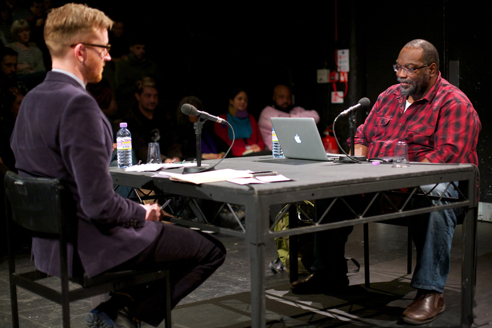 Fred Moten and Barry Esson sit across a table from each other in discussion