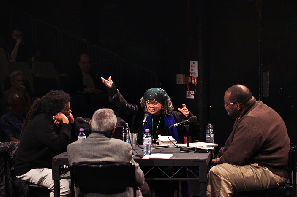 Three folks sit around a table looking at Sonia Sanchez as she gesticulates