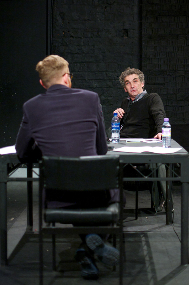 Howard Slater is in conversation at a table with Barry Esson in a black room 