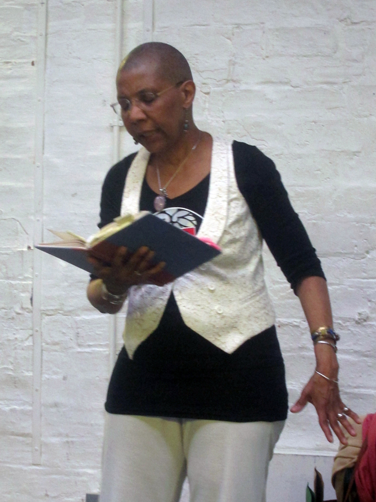 M. NourbSe Philips holds a book and reads a poem by a white brick wall