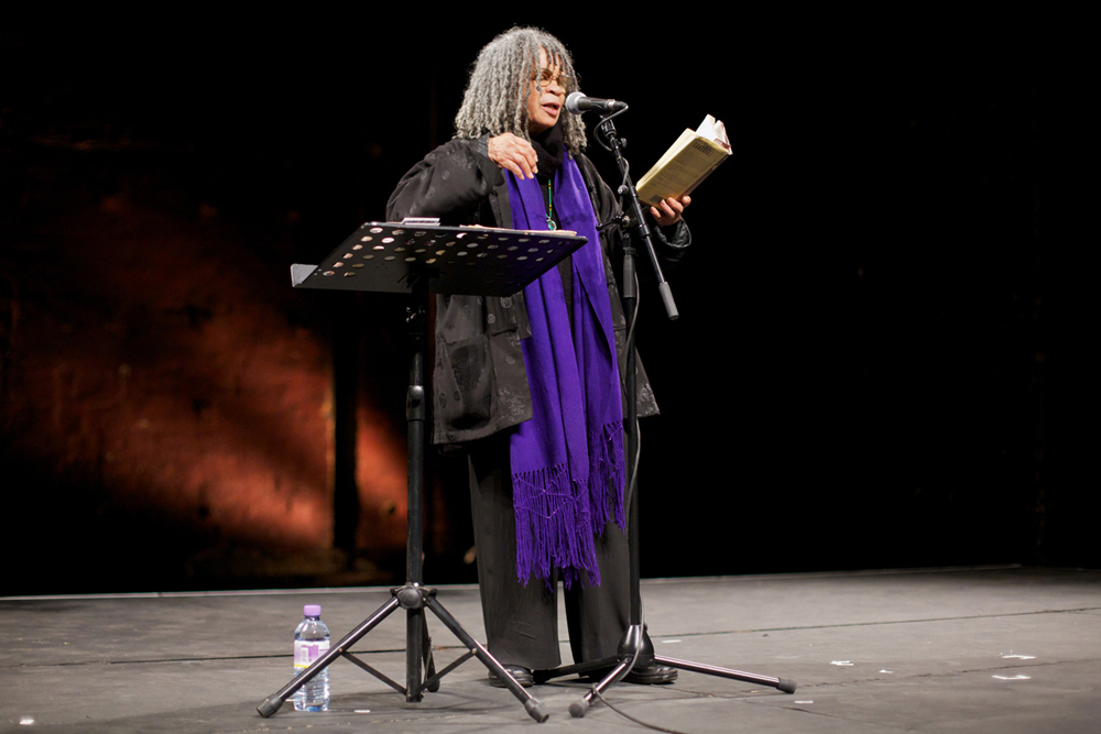 Sonia Sanchez gesticulates as she reads poetry. She wears a purple scarf