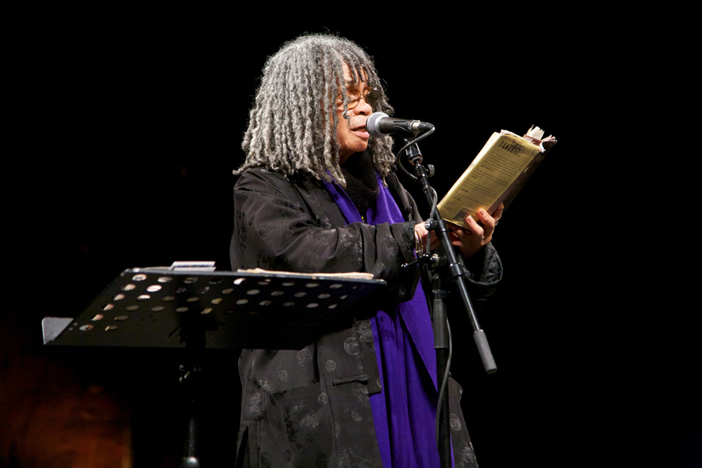 Sonia Sanchez holds a book she reads poetry from it. She wears a purple scarf