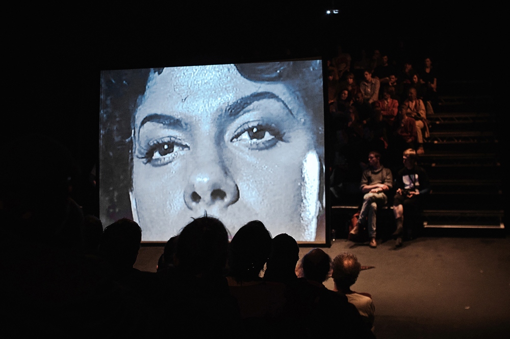 A film plays on a screen, on which there is a B&W close up of a woman's face
