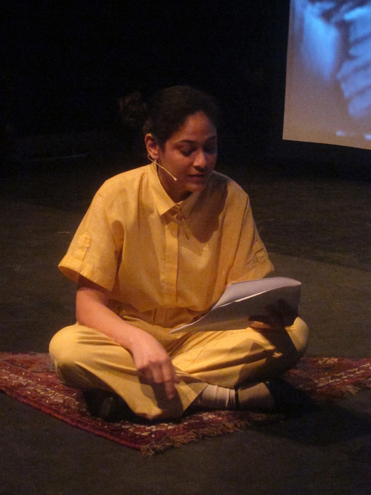 Teresa Maria Diaz Nerio in a yellow jumpsuit reads aloud whist crossed legged