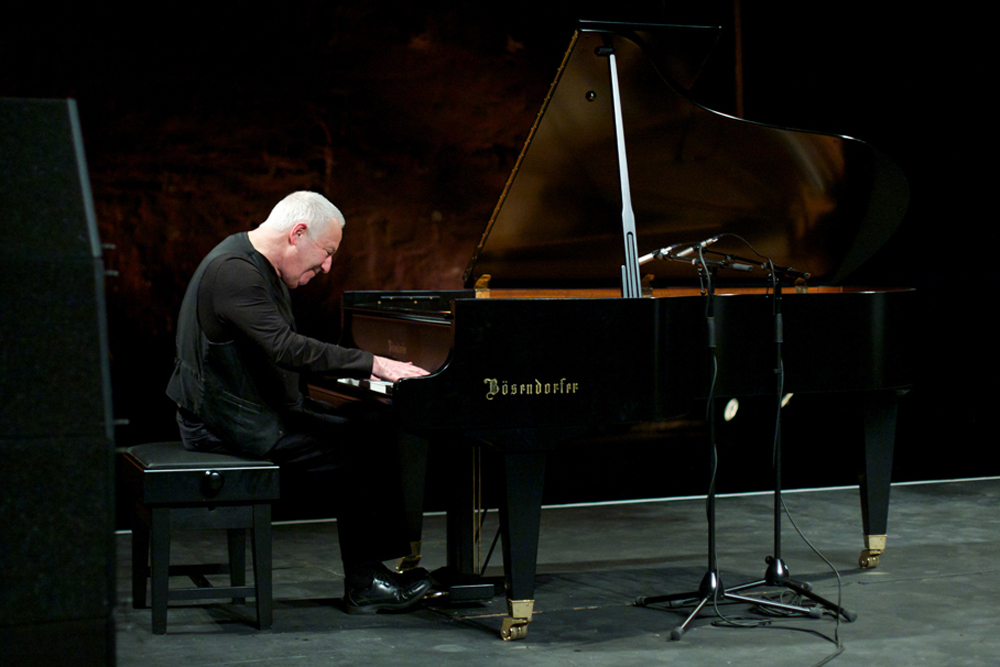 John Tilbury plays a grand piano in a dark theatre space