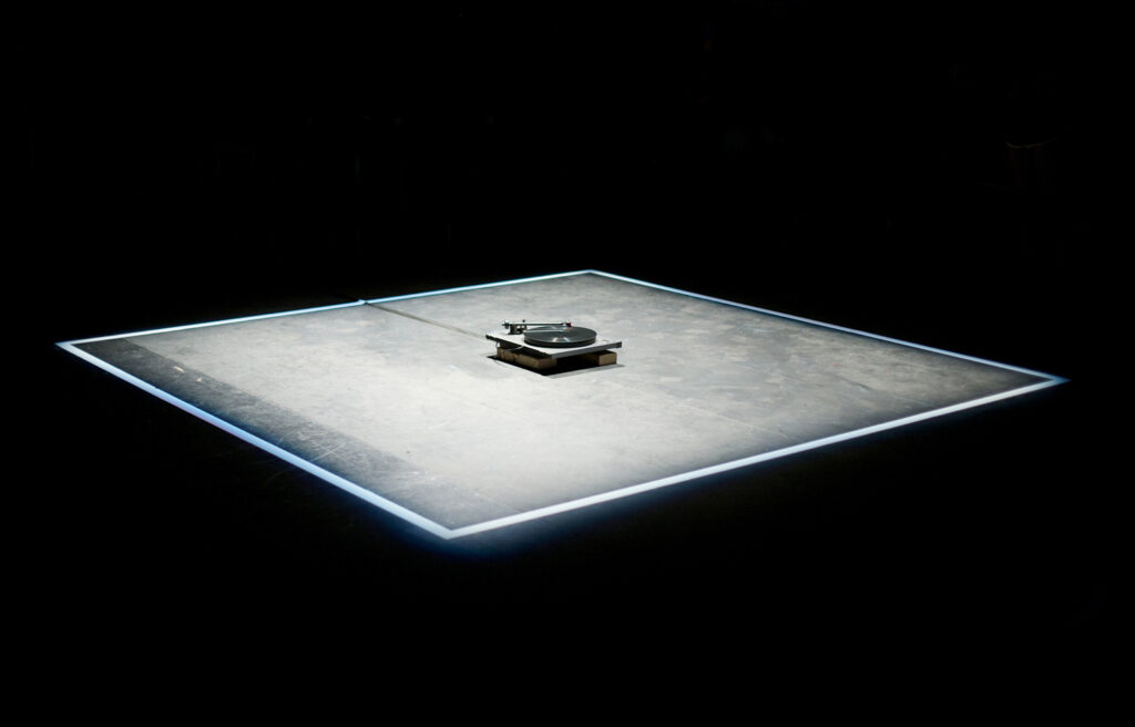 A square of light is on the floor in a dark theatre, a turntable in the centre