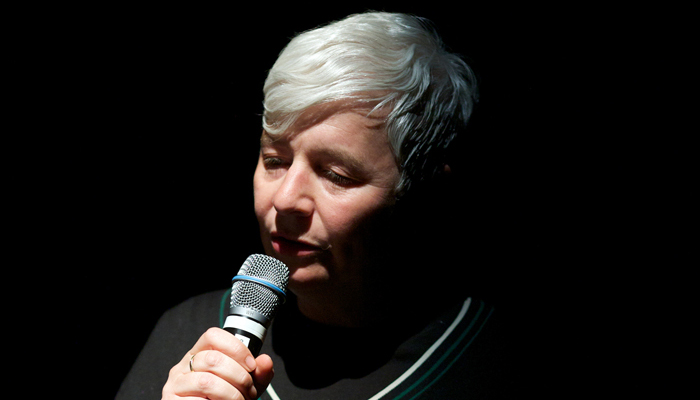 Renate Lorenz speaking on a microphone in a dark performance space