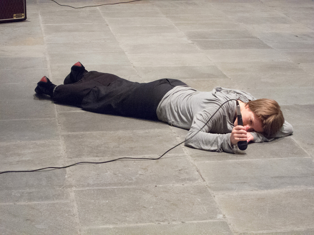 a participant lies on the gallery floor face down and hits the floor with a mic