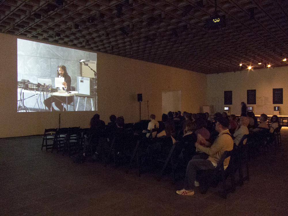 An audience sit in rows in a dark gallery watch a screen showing Rene Gabri