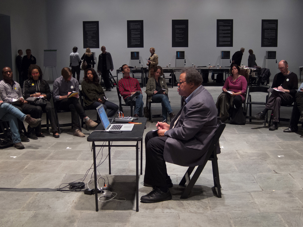 George Lewis sits at a small desk with laptop in the middle of a circle of folk
