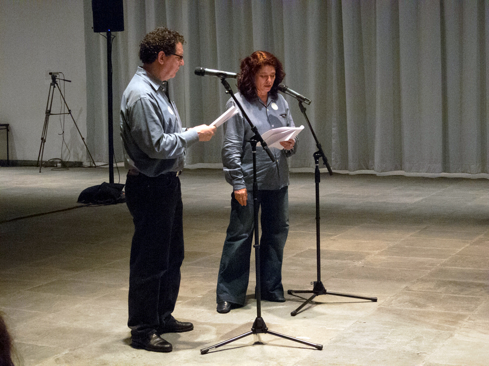 Two Participants stand and speak at microphones whilst reading from scripts