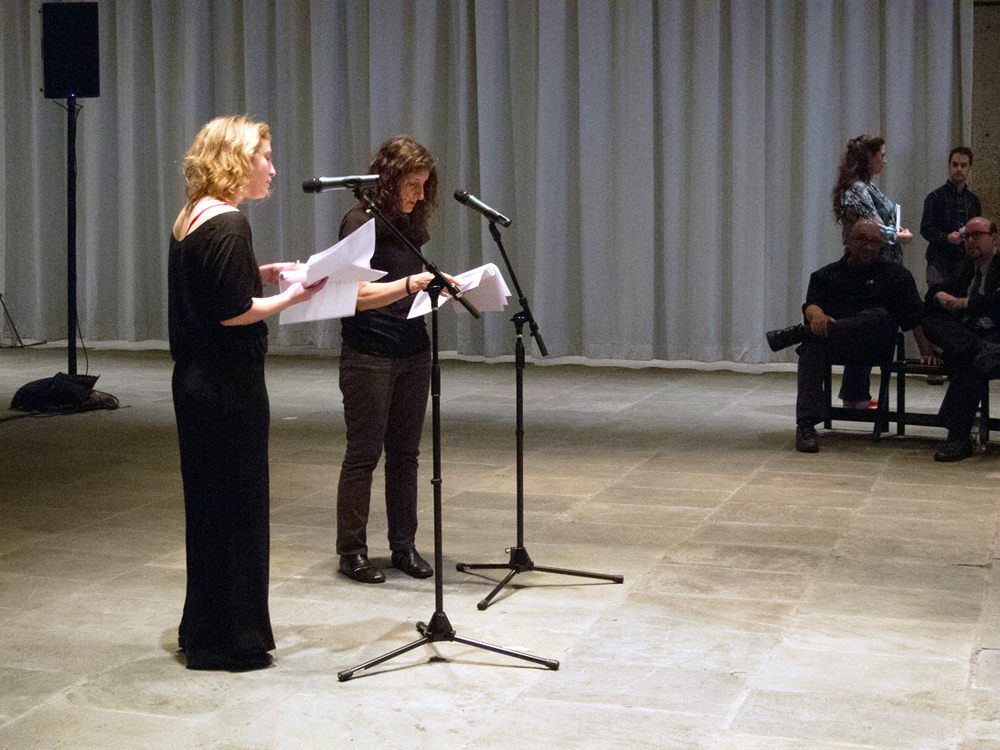 Two Participants stand and speak at microphones whilst reading from scripts
