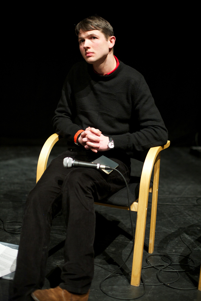 Luke Fowler seated looks out whilst clasping his hands