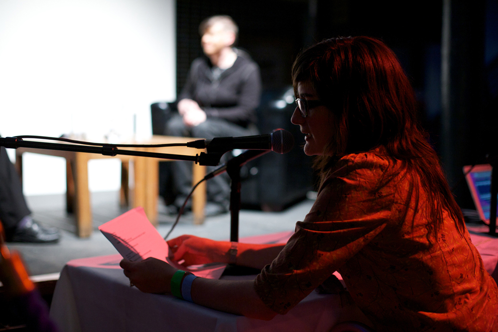 In shadow in the foreground a woman with long hair and glasses reads into a mic
