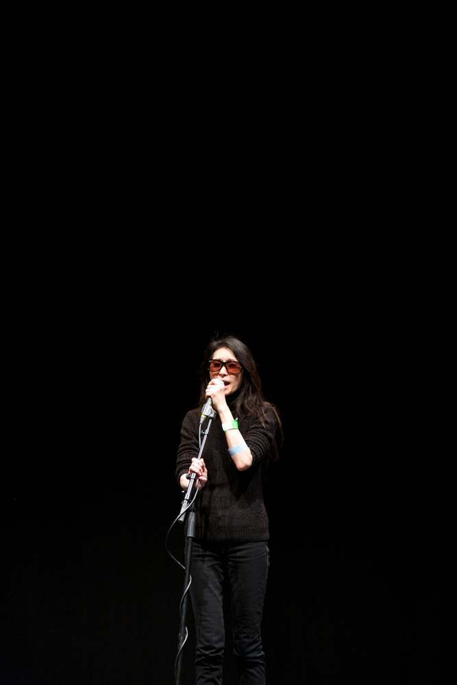 A long shot of Junko in sunglasses and dark clothes on a stage singing
