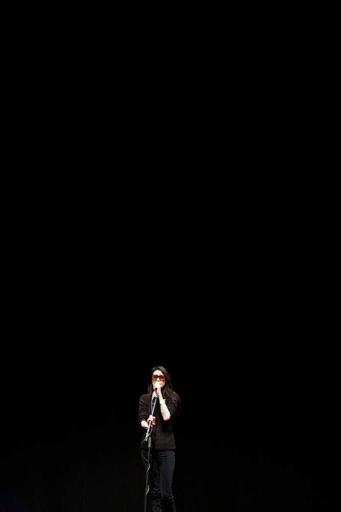 A long shot of Junko in sunglasses and dark clothes on a stage singing