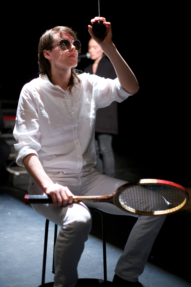 Malin Arnell in white and sunglasses holds a tennis racket and suspended ball