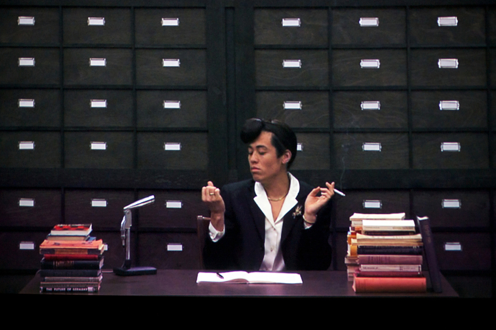 An actor playing a reporter at a trial sits in front of a drawered wall