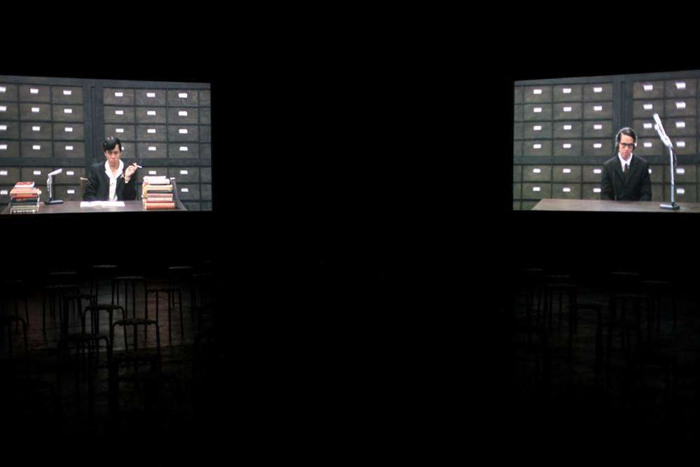Two screens with video of two characters are hung in a dark space