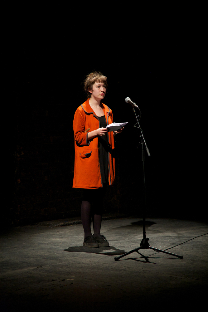 A cast member in an orange coat addresses the audience
