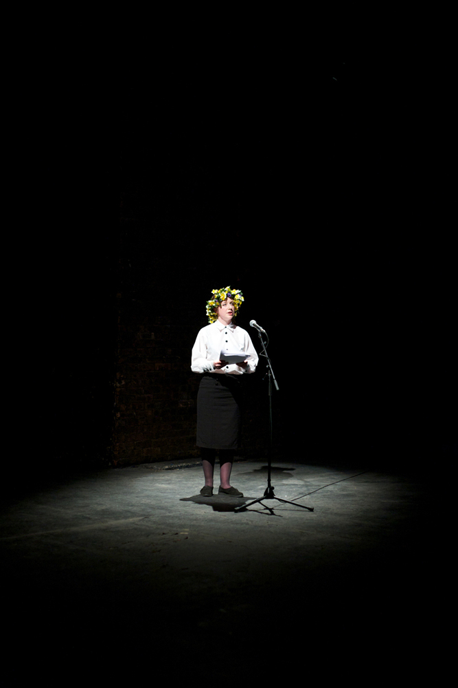 A spotlit female character in a floral headdress addresses the audience