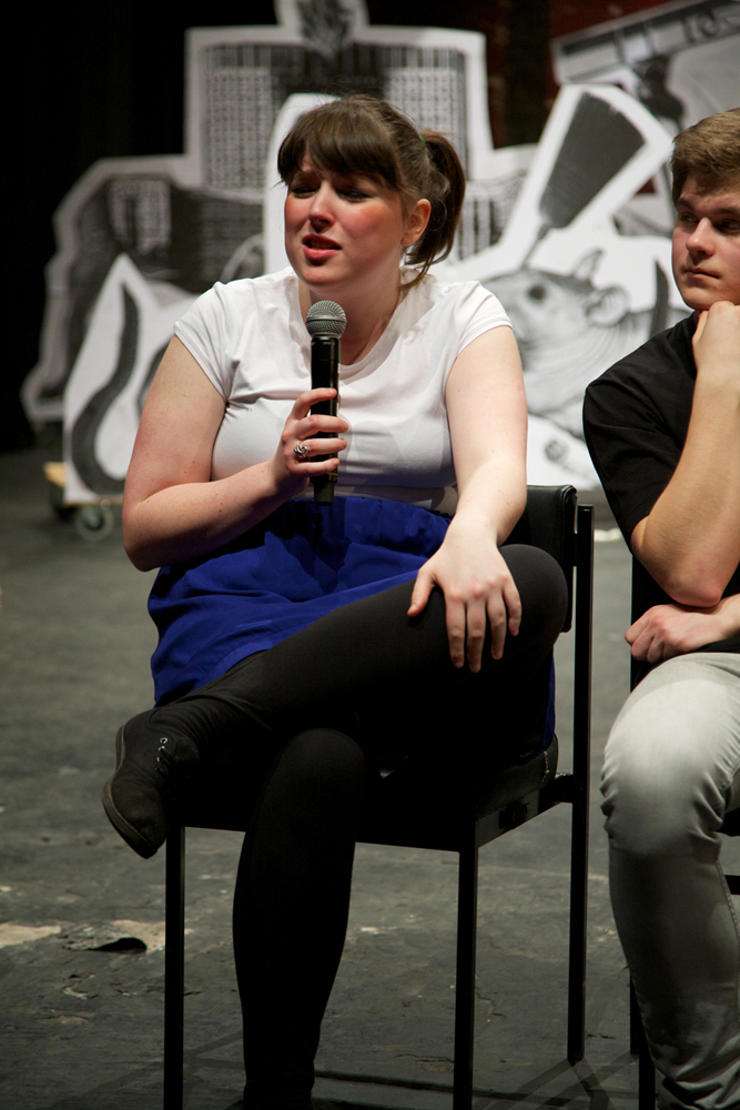 A cast member talks on a microphone whilst seated in front of the stage