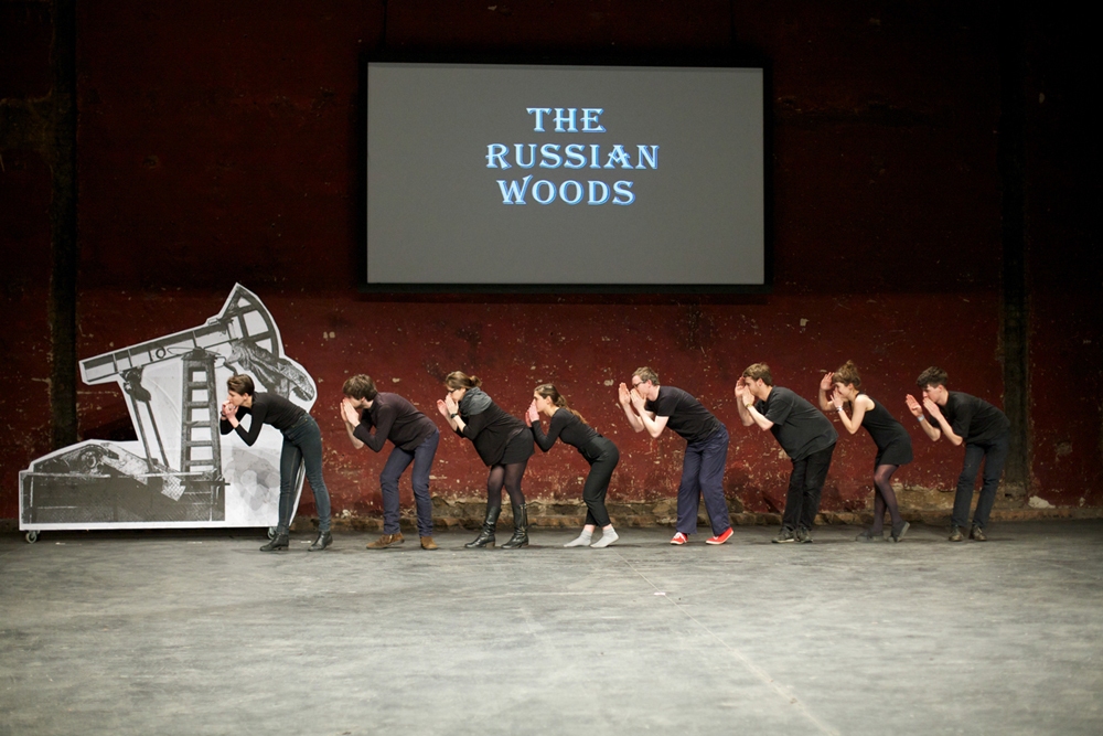 An ensemble of cast bend over in a line, all dressed in black