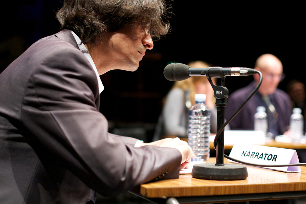 A participant leans into a mic - a sign saying narrator is on their desk