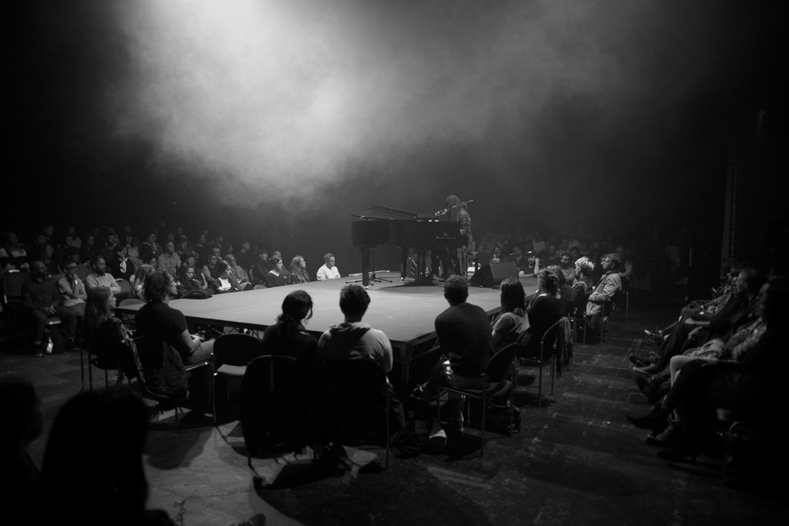 M Lamar on piano on a central stage surrounded by seated audience