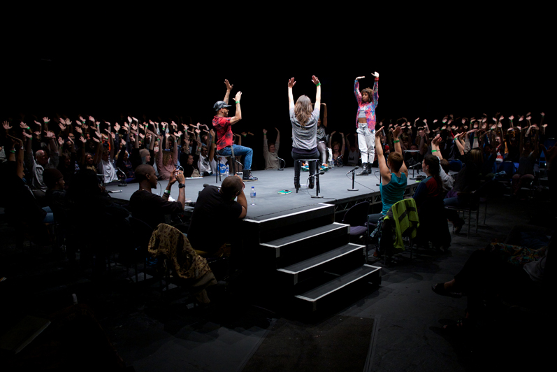 The audience and the folks on the stage raise their hands in the air