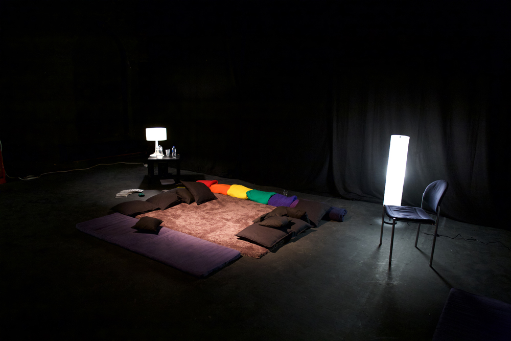 In a dark room, coloured blankets and cushions are arranged 