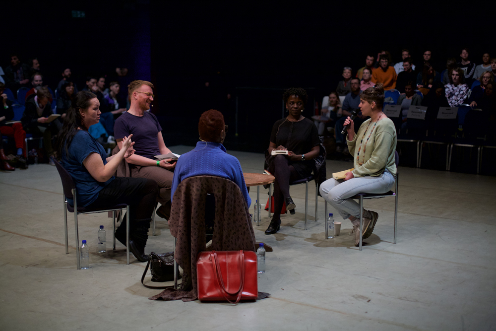 Five people sit in a group in the middle of a space, in discussion