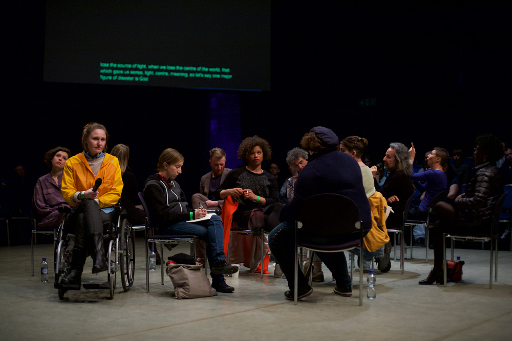 A group of people sit randomly in the centre of a room