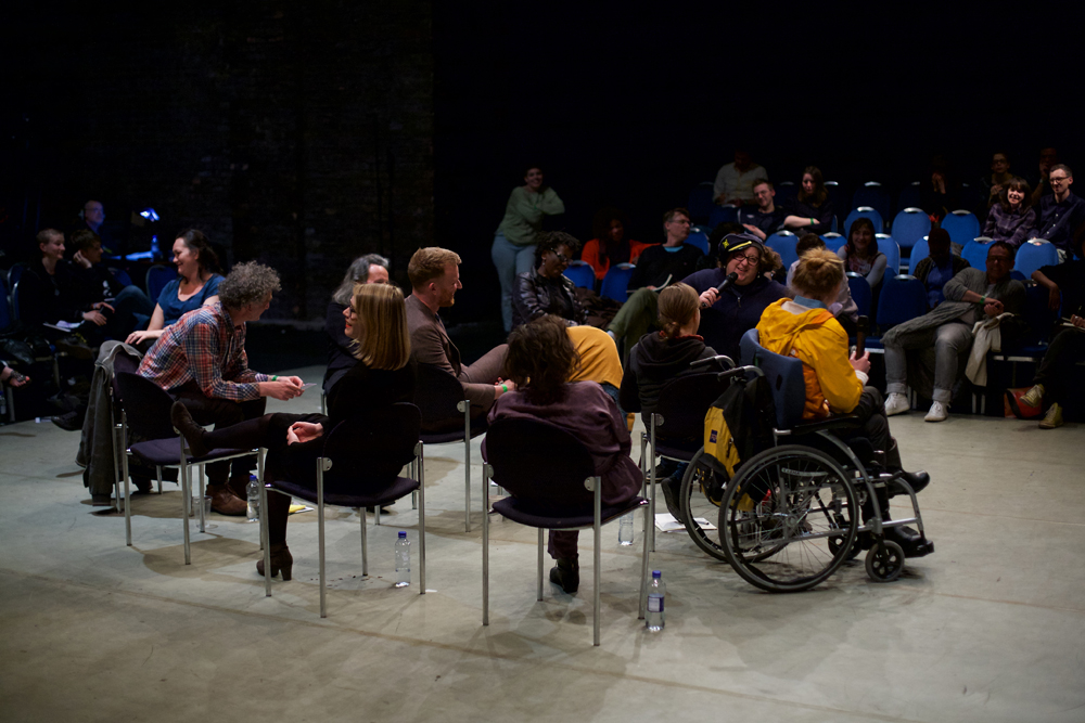 A group of people sit randomly in the centre of a room