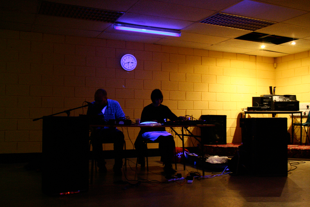 By blacklight in front of a yellow wall two silhouetted people perform