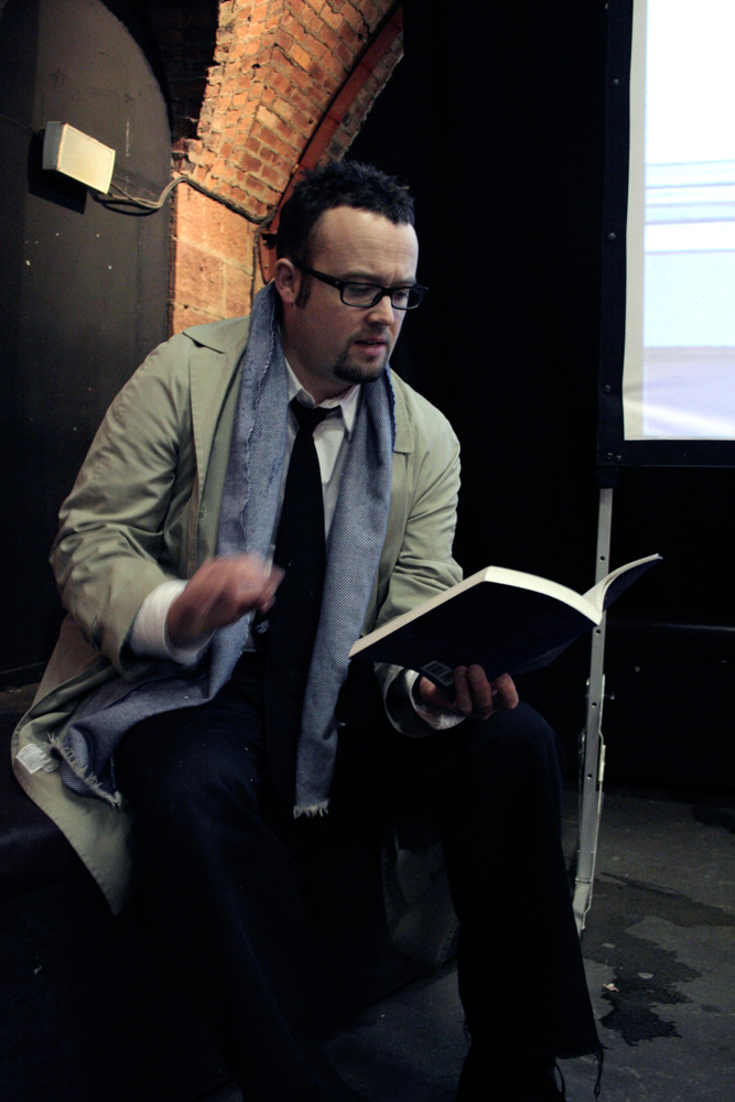 Simon Morris in suit and mac reads from a book