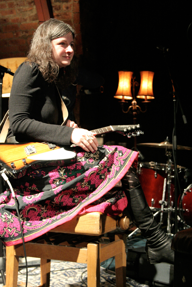 Erica Elder sits with a guitar on her lap smiling in a floral skirt