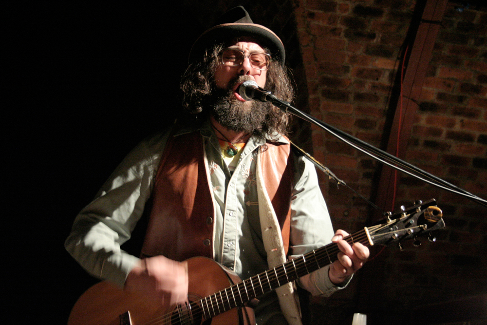 a Bearded and hatted Matt Valentine plays guitar and sings 