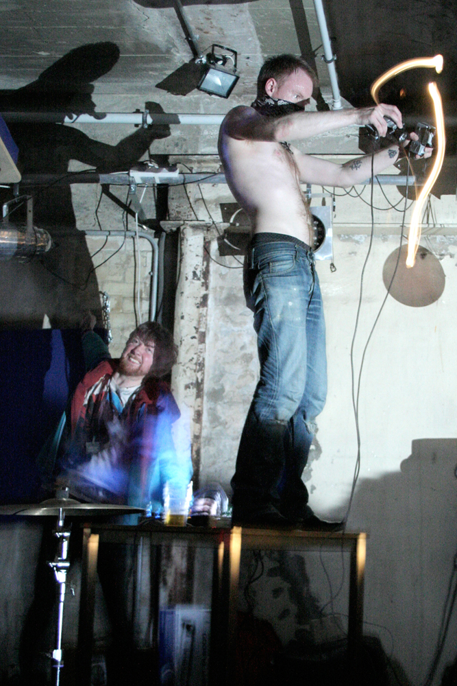 A bare chested man with a bandana of their face stands on a table
