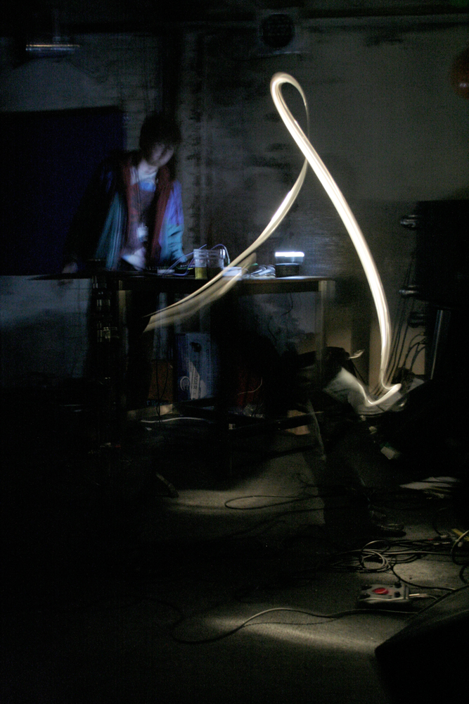 A performer in a dark space plays electronics, a light streak in the foreground