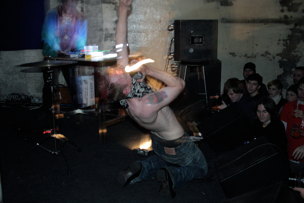 A bare chested man with a bandana of their face screams whilst moving a torch